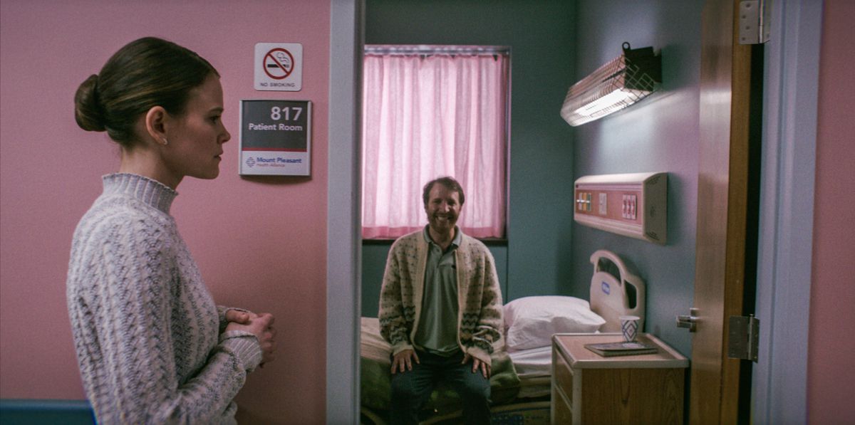 Rose (Sosie Bacon) in piedi fuori da una stanza d'ospedale in cui un uomo barbuto con un maglione cardigan siede dritto sul suo letto d'ospedale sorridendo come un maniaco in Smile