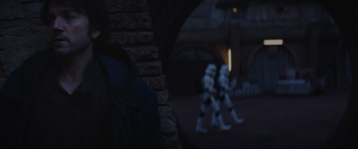 Cassian Andor si nasconde dietro un pilastro di notte mentre gli Stormtroopers camminano per le strade dietro di lui