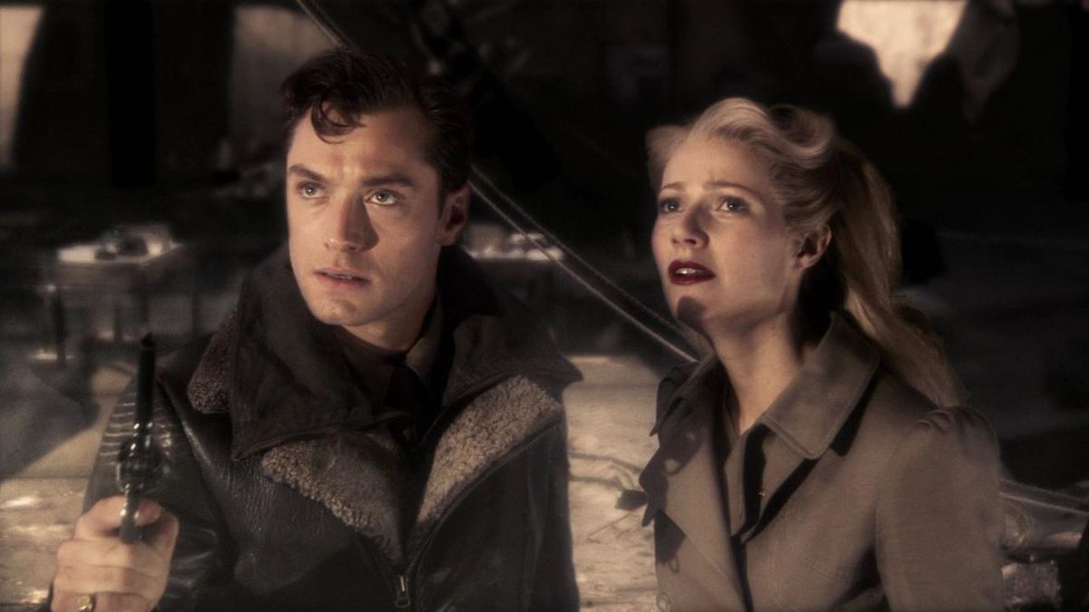 Un uomo (Jude Law) con indosso una giacca da pilota di pelle con in mano una pistola è in piedi accanto a una donna (Gwyneth Paltrow) con una giacca marrone chiaro che fissa qualcosa fuori dallo schermo. 