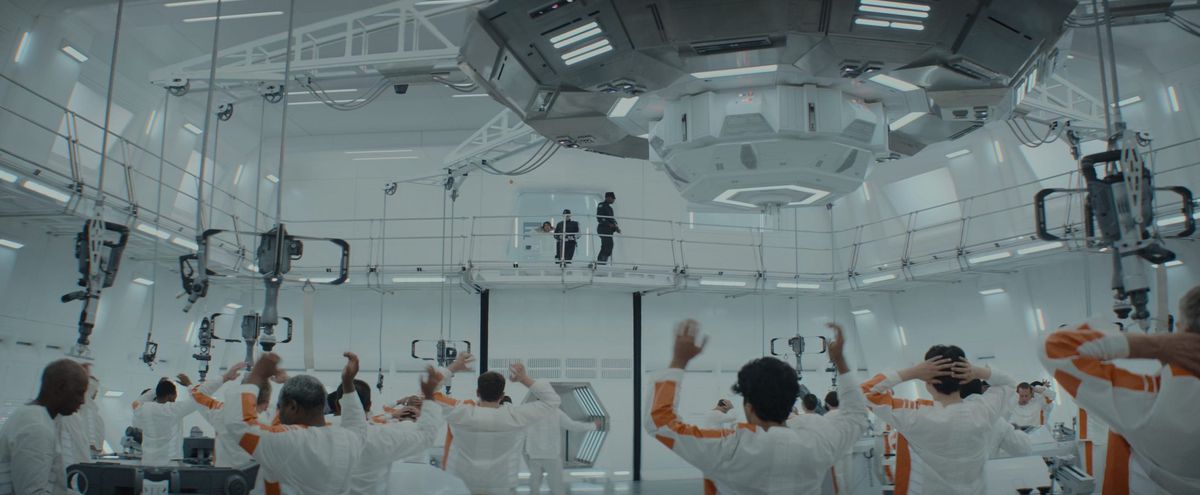 I prigionieri in tute bianche e arancioni invecchiano le mani in aria sotto giganteschi macchinari mentre un nuovo prigioniero viene scortato al piano dell'area di lavoro tramite una passerella di ascensore ad Andor