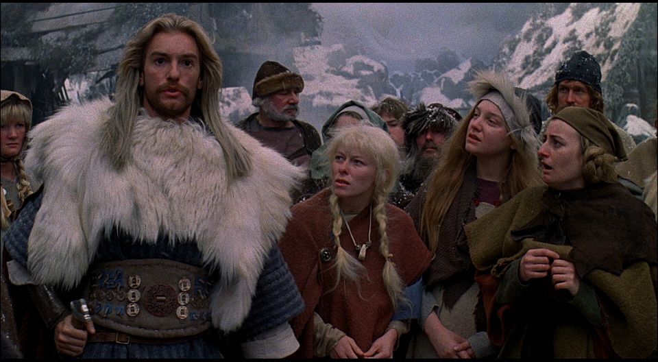 Un gruppo di abitanti di un villaggio vichingo si trova tra montagne innevate, con un uomo alto con pelliccia bianca e lunghi capelli biondi, in Erik il Vichingo.