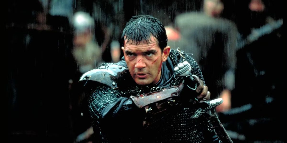 Un uomo con cicatrici visibili sul viso (Antonio Banderas) estrae la spada dal fodero mentre è bagnato dalla pioggia.