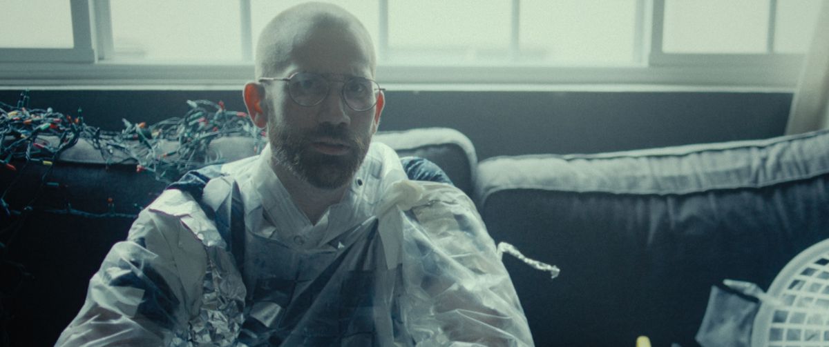 John Daniels (sceneggiatore-regista Aaron Moorhead) siede su un divano sudicio in una stanza poco illuminata, indossando un abito sciatto fatto di nastro adesivo, carta stagnola e teli di plastica, in Qualcosa nella sporcizia