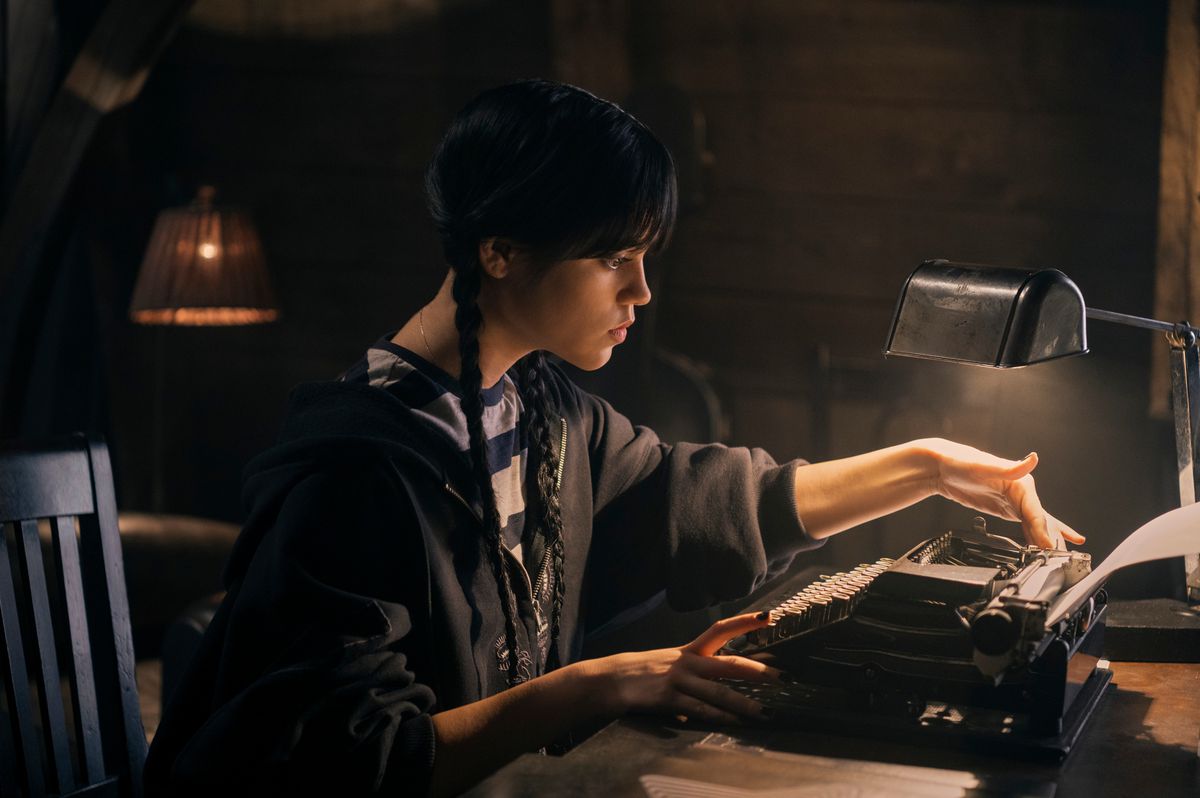 mercoledì, una ragazza dai capelli scuri con i capelli raccolti in trecce, china su una macchina da scrivere in una stanza scarsamente illuminata 