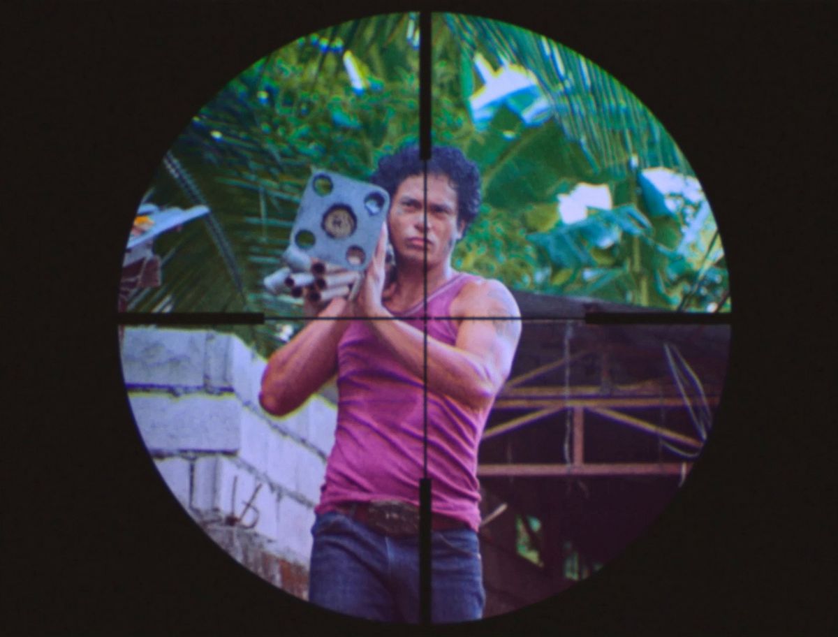 Un uomo filippino con i capelli ricci, che indossa una maglietta muscolare rosa, è visto in un video a bassa risoluzione attraverso la vista di un cecchino in una sequenza di film nel film di Leonor Will Never Die