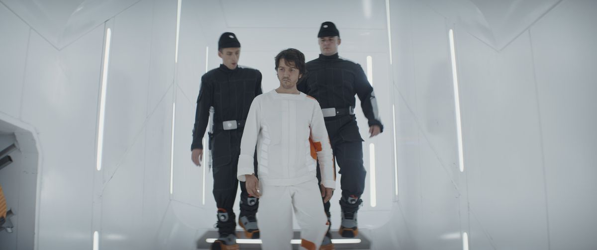 Due guardie che indossano uniformi imperiali grigie e gigantesche scarpe da ginnastica isolanti conducono Cassian Andor, in un body bianco con finiture arancioni, giù per le scale di una prigione tutta bianca