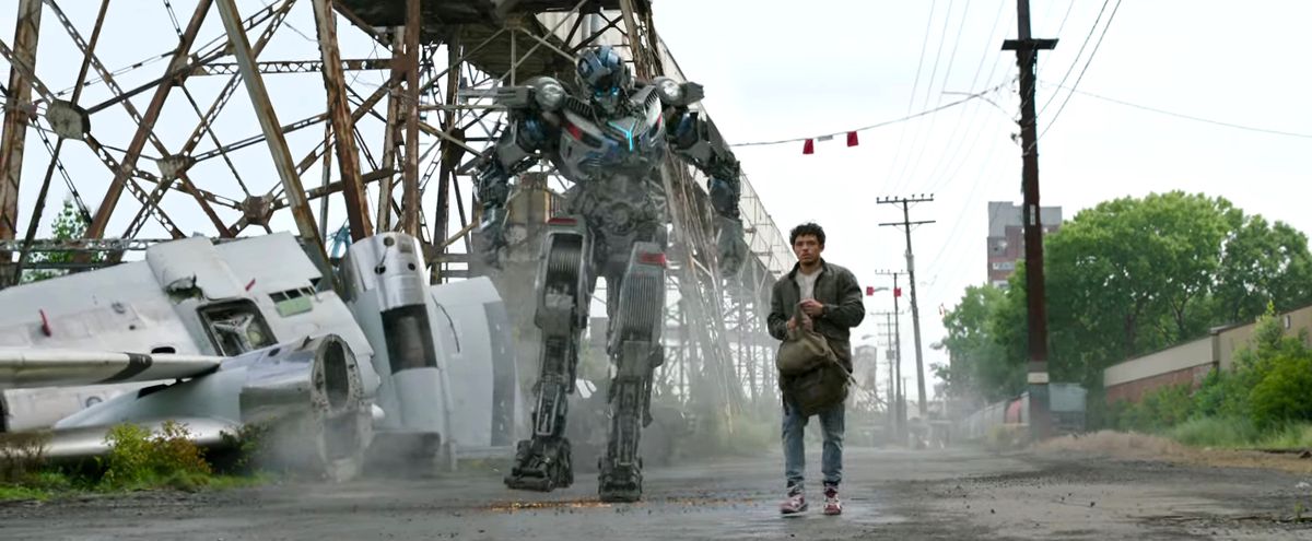 Mirage e un umano ispanico che trasportano uno zaino camminano lungo una strada polverosa in Transformers: Rise of the Beasts