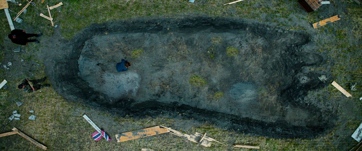 Una foto dall'alto verso il basso di un'immensa impronta di troll conficcata in profondità in un campo erboso, con minuscole figure umane che la esaminano di lato, nel Troll del 2022