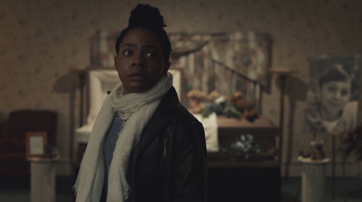 Monique (Gabby Beans), una donna dalla pelle scura con una giacca nera e una sciarpa bianca, si trova in una stanza buia con una bara aperta dietro di lei e sembra allarmata per qualcosa fuori campo in The Harbinger