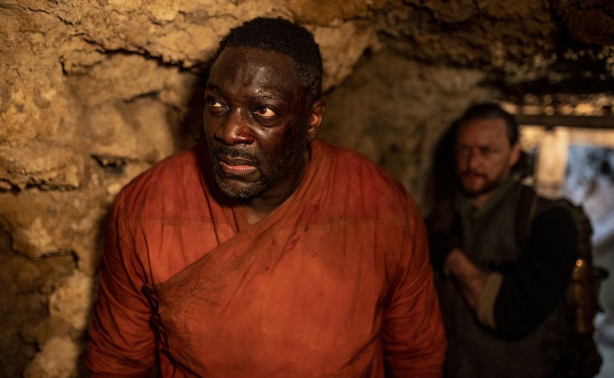 Il comandante Ogunwe (Adewale Akinnuoye-Agbaje) in piedi in una caverna che guarda qualcosa con Asriel (James McAvoy) dietro di lui in un fotogramma della stagione 3 di His Dark Materials