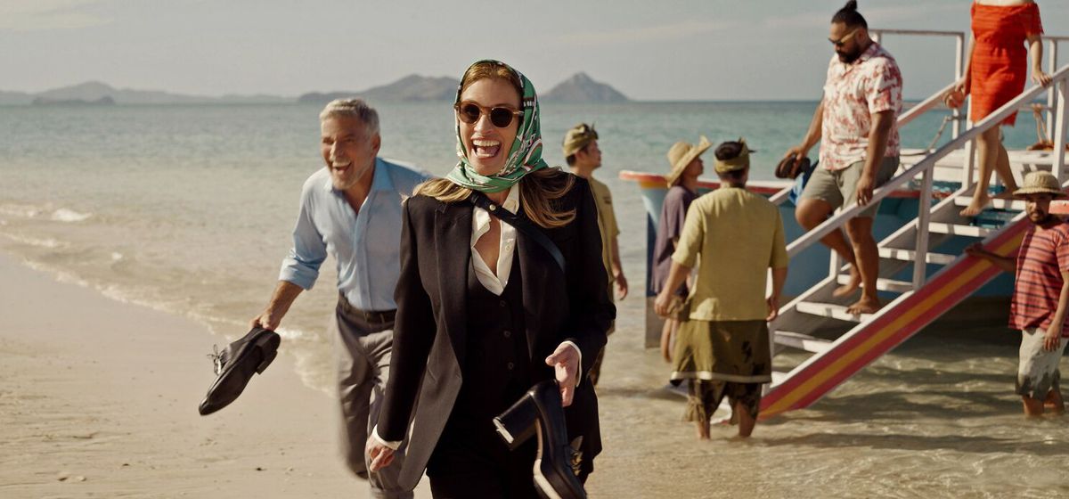 Un uomo (George Clooney) e una donna che indossa una sciarpa sopra la testa (Julia Roberts) ridono mentre sbarcano da una barca su una spiaggia e tengono le scarpe in mano.