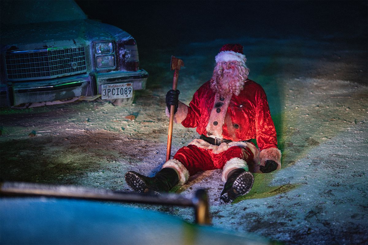 Un uomo vestito da Babbo Natale coperto di sangue (Abraham Benrubi) impugna un'ascia mentre è seduto in un campo innevato accanto a un'auto parcheggiata.