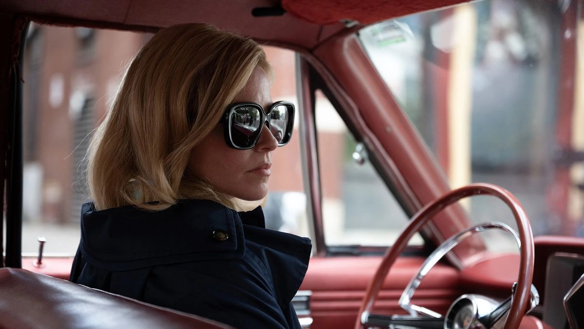 Una donna che indossa un cappotto blu e occhiali (Elizabeth Banks) siede al volante di un'automobile rossa.