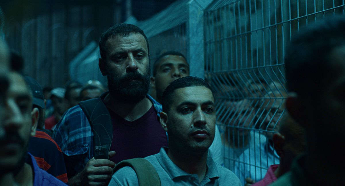 Un uomo con la barba (Ali Suliman) si trova in una fila affollata di persone lungo una recinzione collegata a una catena.