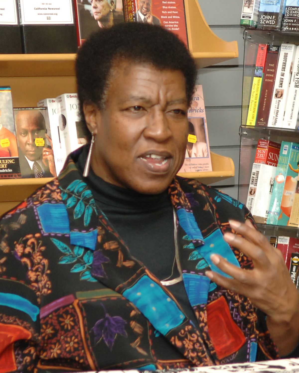 Octavia E. Butler negli anni '90, che indossa una camicia fantasia con disegni di foglie d'albero e lunghi orecchini di metallo, viene colta a metà frase durante una lettura in una libreria