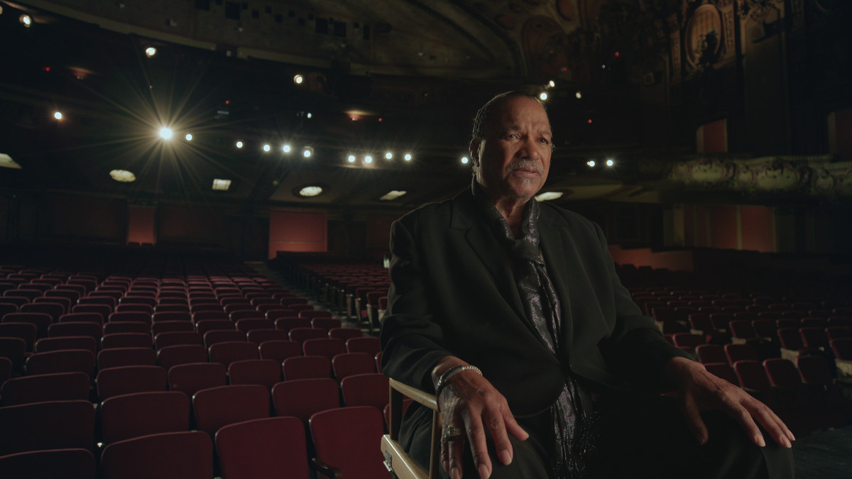 Billy Dee Williams siede su una sedia da regista su un palcoscenico teatrale, con la schiena rivolta verso le sedie vuote del pubblico