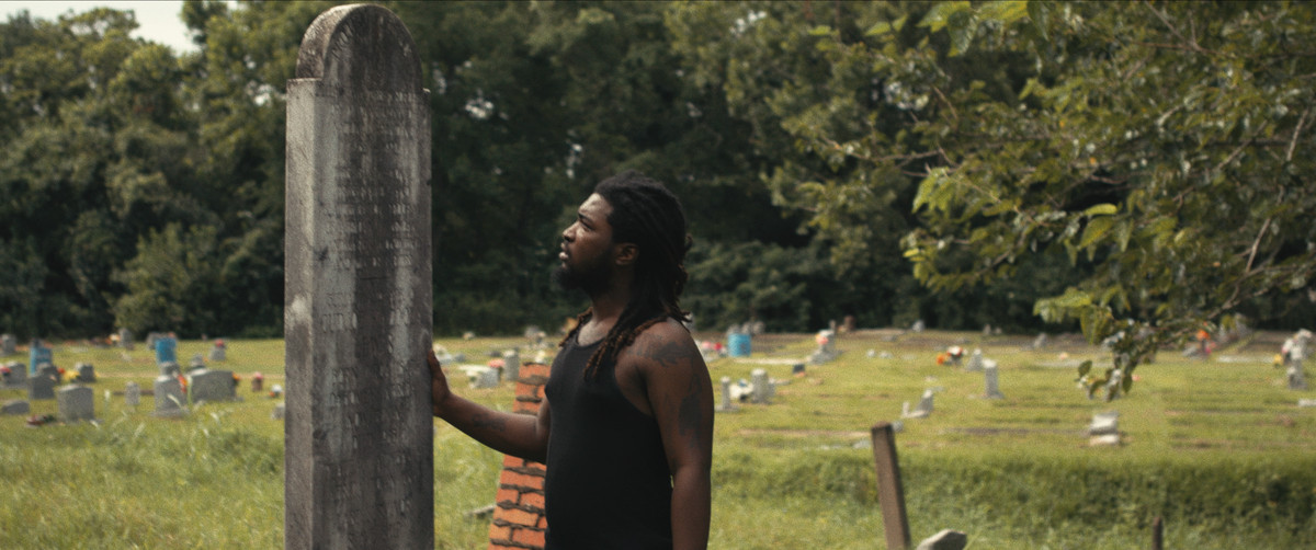 Un uomo di colore con i dreadlocks e una canottiera nera si trova in un cimitero e posa una mano su un'imponente lapide su cui sono incisi dei nomi