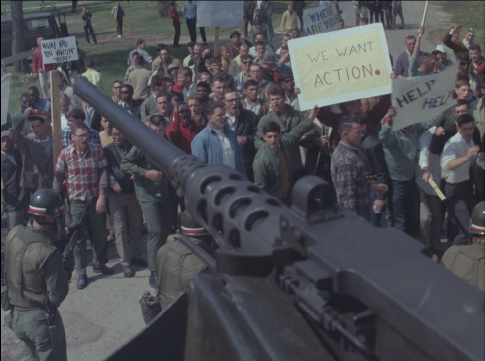 Un cannone da carro armato entra nell'inquadratura sopra una folla di manifestanti in scena, uno dei quali tiene un cartello con la scritta 