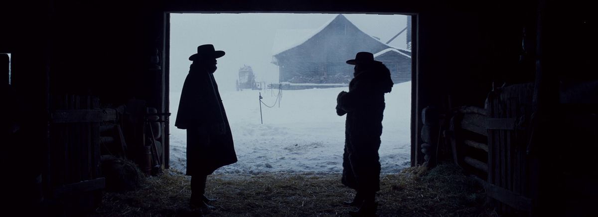 Due uomini in cappotti invernali e cappelli da cowboy si trovano all'interno di un fienile con un altro edificio visibile in lontananza e una tempesta di neve all'esterno.