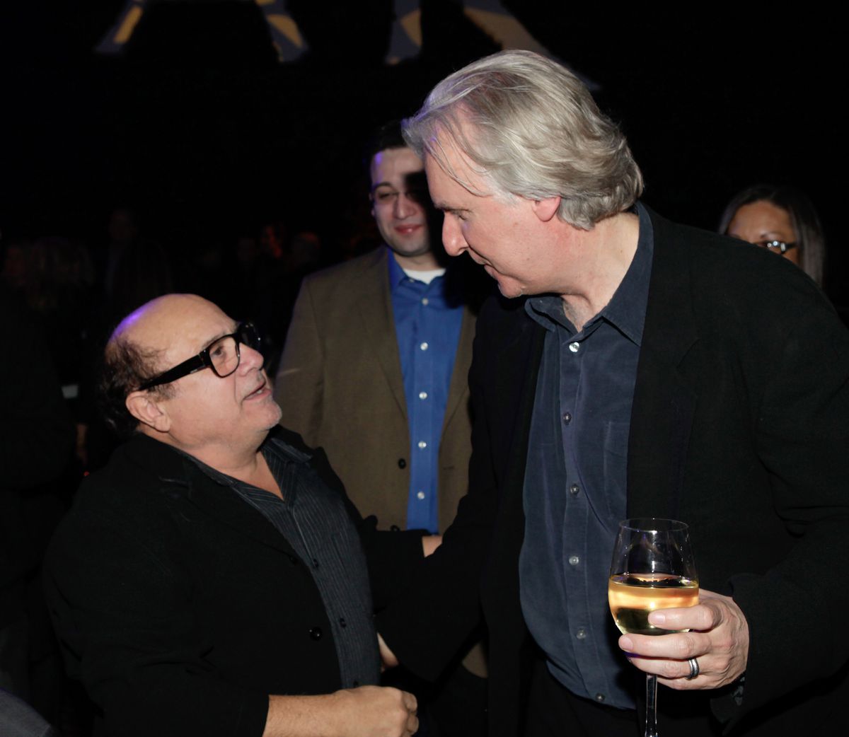 Danny Devito alza lo sguardo per parlare con il regista James Cameron, che ha in mano un bicchiere di vino bianco all'after party di Avatar