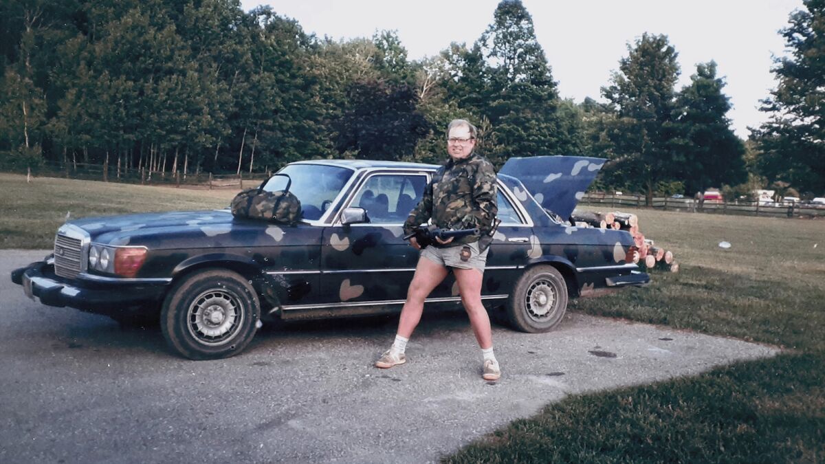 Una vecchia foto degli anni '80 mostra un uomo in mimetica e pantaloncini corti che attraversa un campo fino a un parcheggio dove è parcheggiata la sua berlina mimetica 