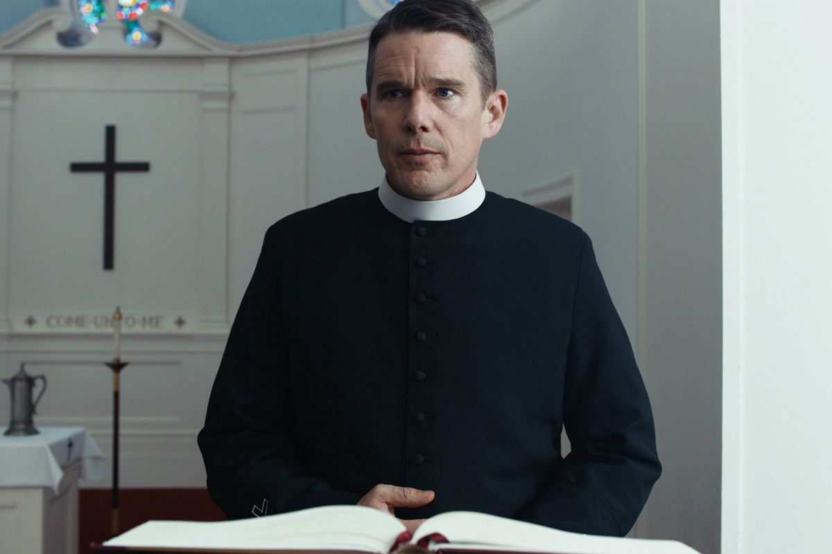Un uomo in abito da prete (Ethan Hawke) è in piedi dietro un leggio con una croce nera visibile sullo sfondo.
