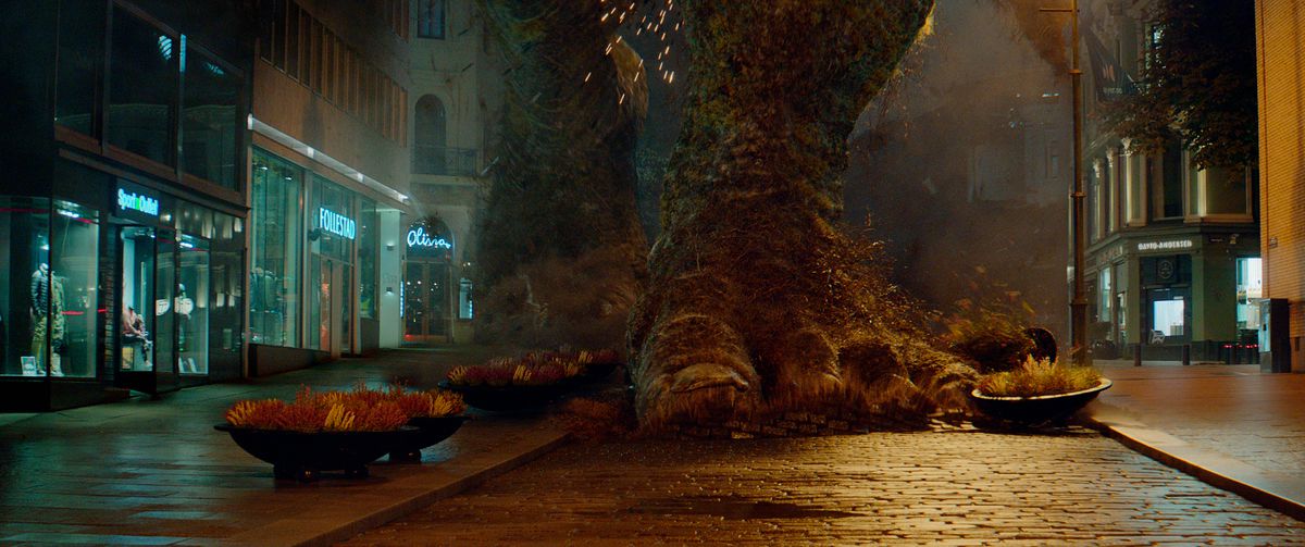 I giganteschi piedi di pietra ricoperti di muschio di un troll calpestano fioriere nel mezzo di una strada cittadina notturna nel Troll del 2022