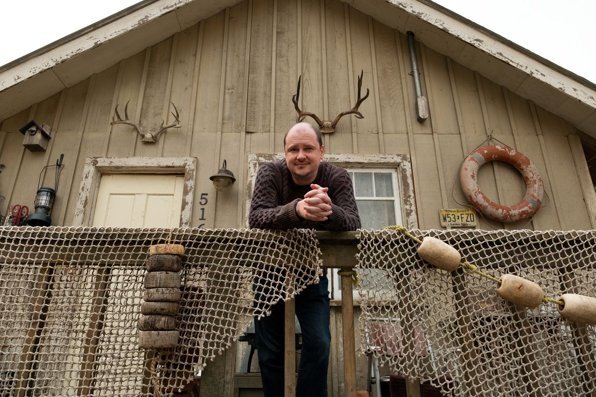Mike Flanagan si trova di fronte a una casa di legno con sopra delle corna e un salvavita.  Si appoggia alla ringhiera.