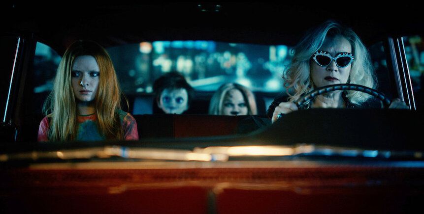 Jennifer Tilly guida un'auto, con un giovane sul sedile del passeggero e due bambole sul sedile posteriore, in Chucky.