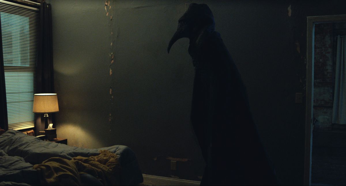 Una figura del medico della peste - alta, scura, mascherata da uccello e ammantata di nero - incombe nell'ombra in una stanza fatiscente con la carta da parati che si stacca dalle pareti in The Harbinger