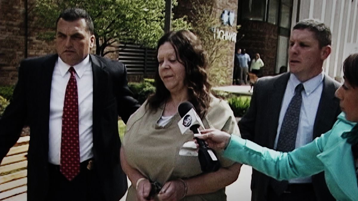 Una donna in camicia marrone chiaro e manette affiancata da due uomini corpulenti in giacca e cravatta viene intervistata da una donna in giacca turchese con in mano un microfono.