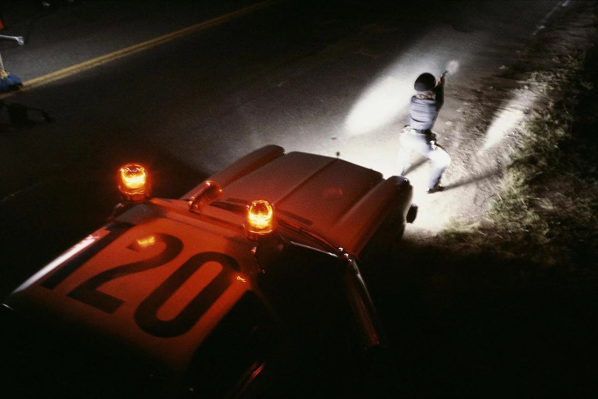 Una ripresa dall'alto di un agente di polizia che indossa un berretto accovacciato davanti a un'auto della polizia di notte, con la schiena illuminata dai fari dell'auto mentre punta la pistola contro qualcosa fuori campo.