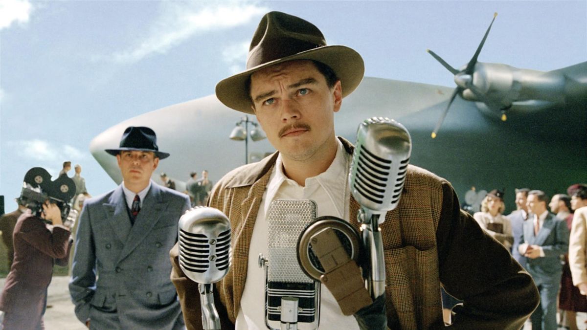 Un uomo con un cappello marrone e folti baffi (Leonardo DiCaprio) si trova di fronte a una serie di aste microfoniche con un gigantesco aeroplano cromato visibile dietro di lui.