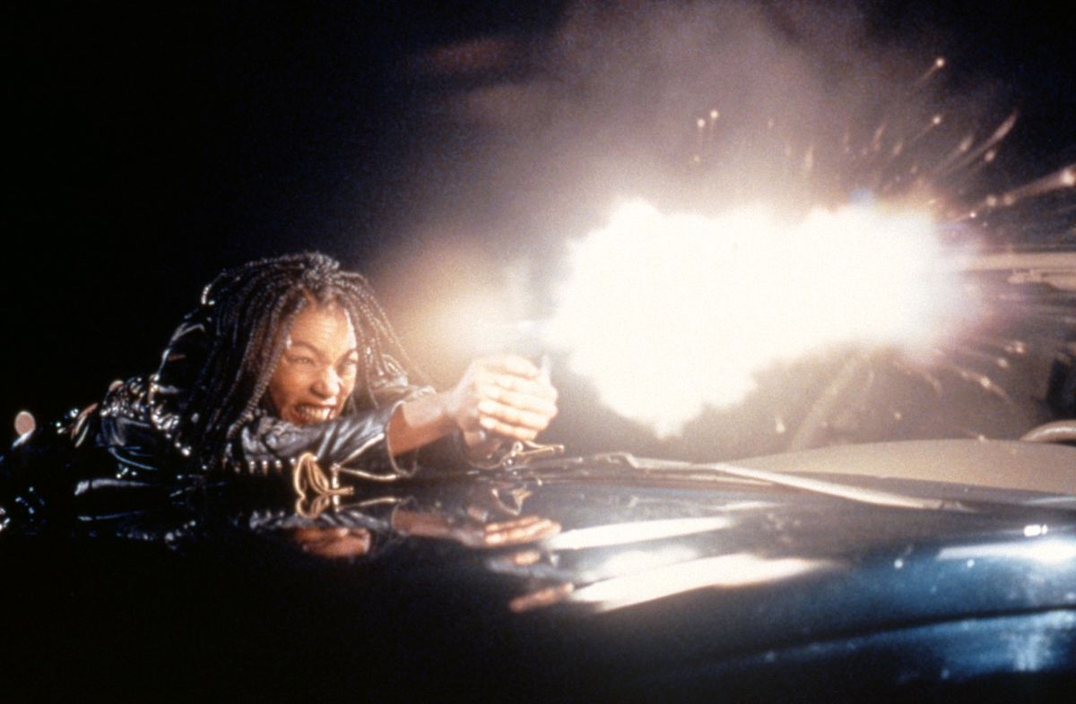 Una donna con una giacca di pelle nera (Angela Bassett) si sporge sopra il cofano di un veicolo scuro e spara con una pistola, un lampo luminoso che illumina sia lei che la superficie del veicolo.