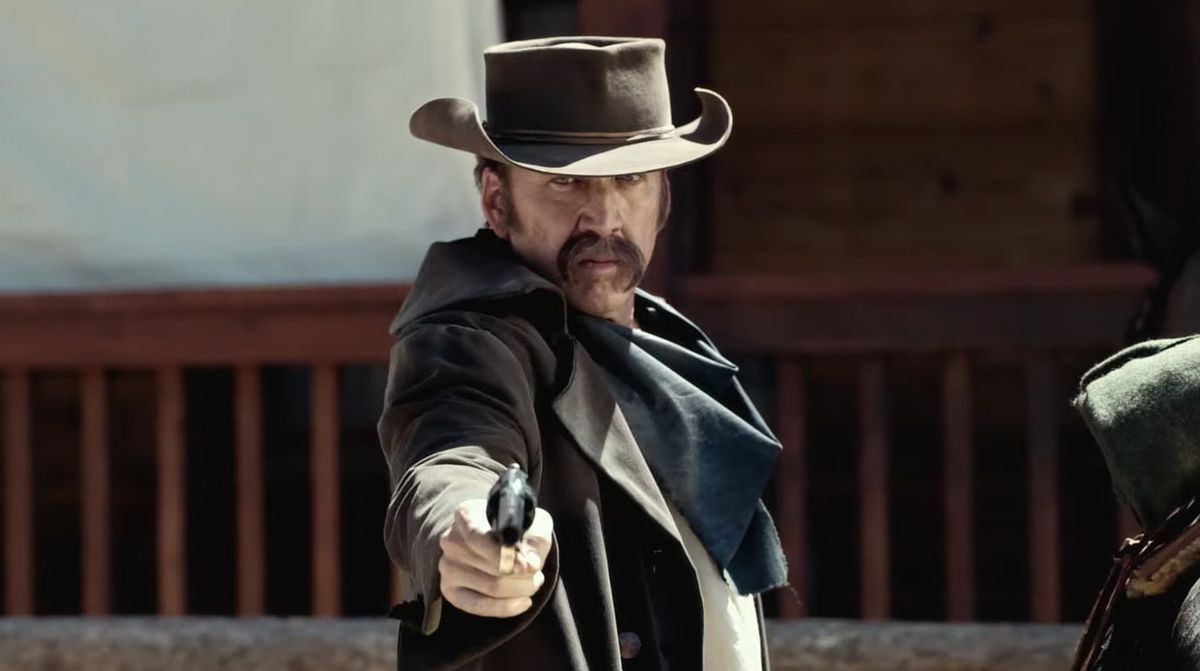 Un uomo con i baffi troppo cresciuti (Nicolas Cage) che indossa un cappello da cowboy e uno spolverino marrone scuro che punta una pistola.