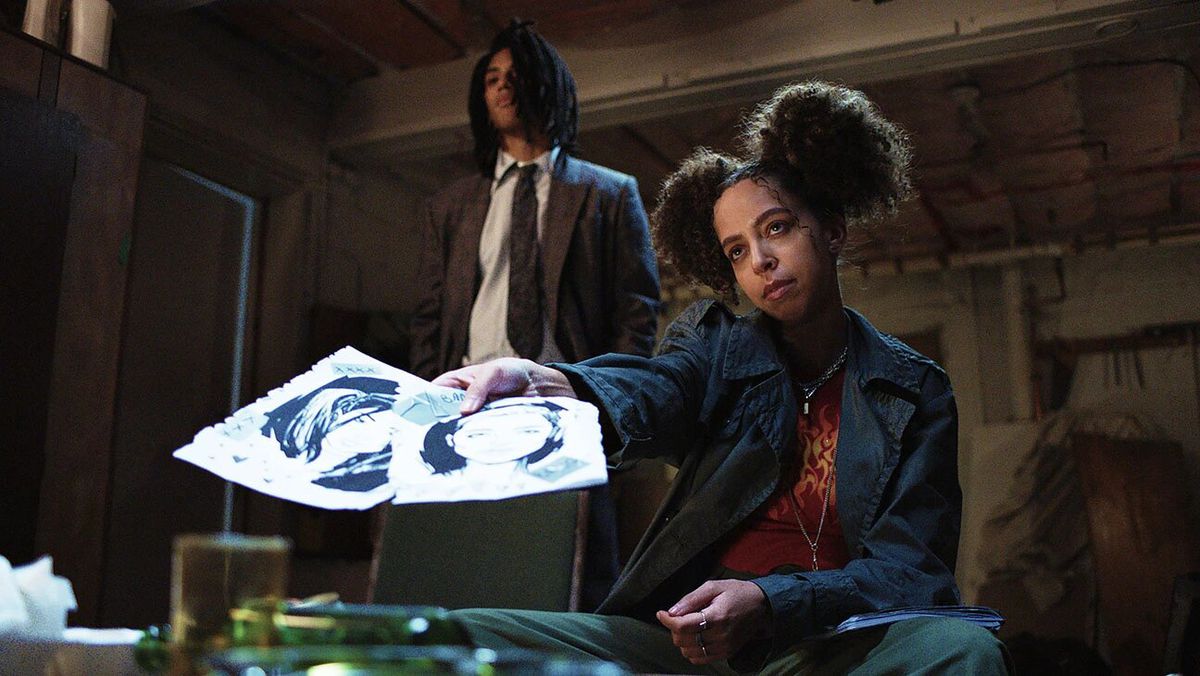 Una donna con i capelli arruffati (Hayley Law) porge due disegni mentre è seduta dietro un tavolo.