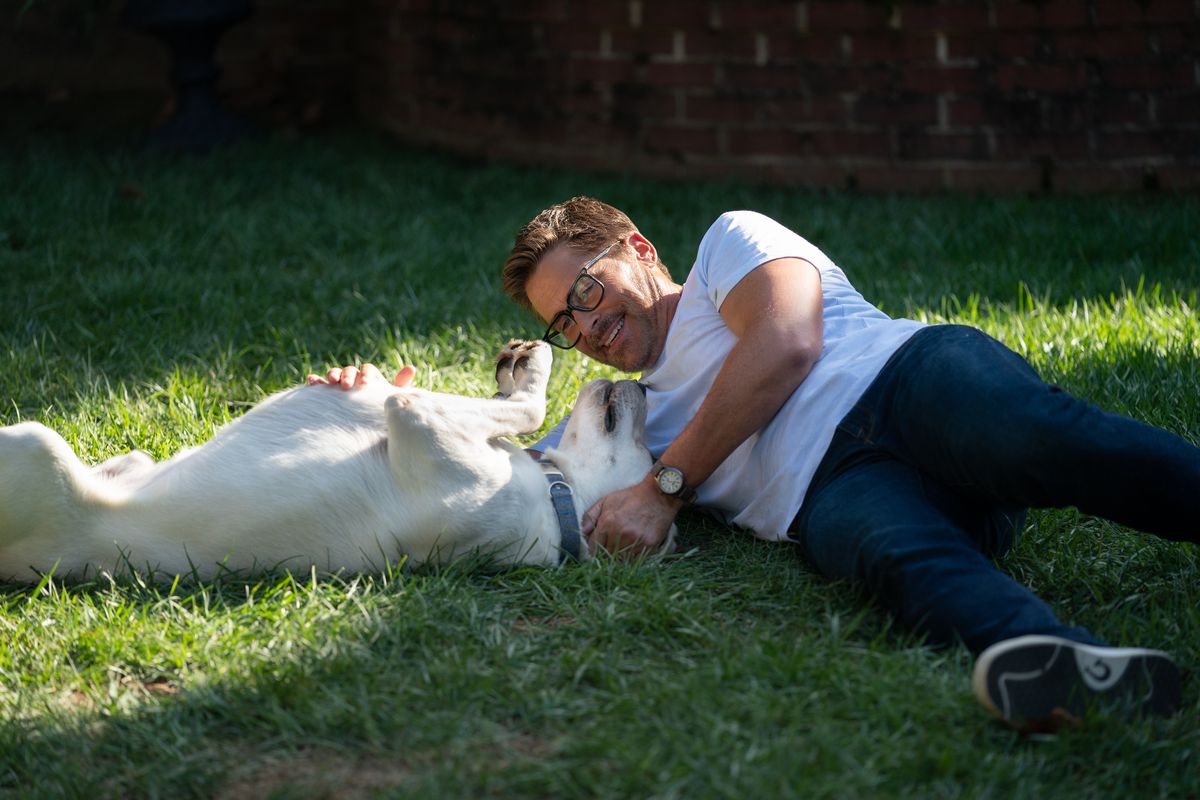 Un uomo (Rob Lowe) con gli occhiali, una maglietta bianca e jeans blu sdraiato su un prato e che gioca con un cane.