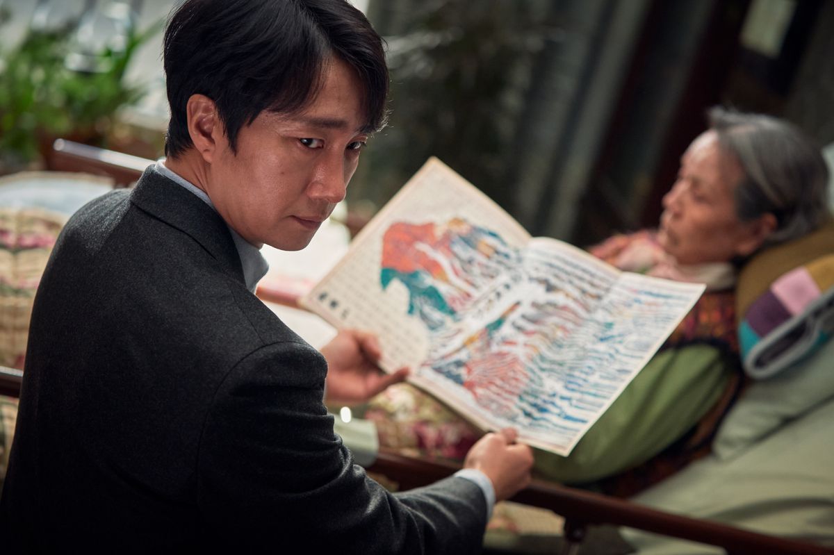 Il detective Hae-jun (Park Hae-il) siede accanto a una donna anziana e apre una colorata pagina simile a una mappa ricoperta di segni indistinti e scritte in Decision To Leave