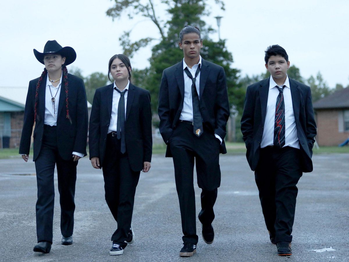 Quattro adolescenti in giacca e cravatta nera che camminano in un parcheggio.