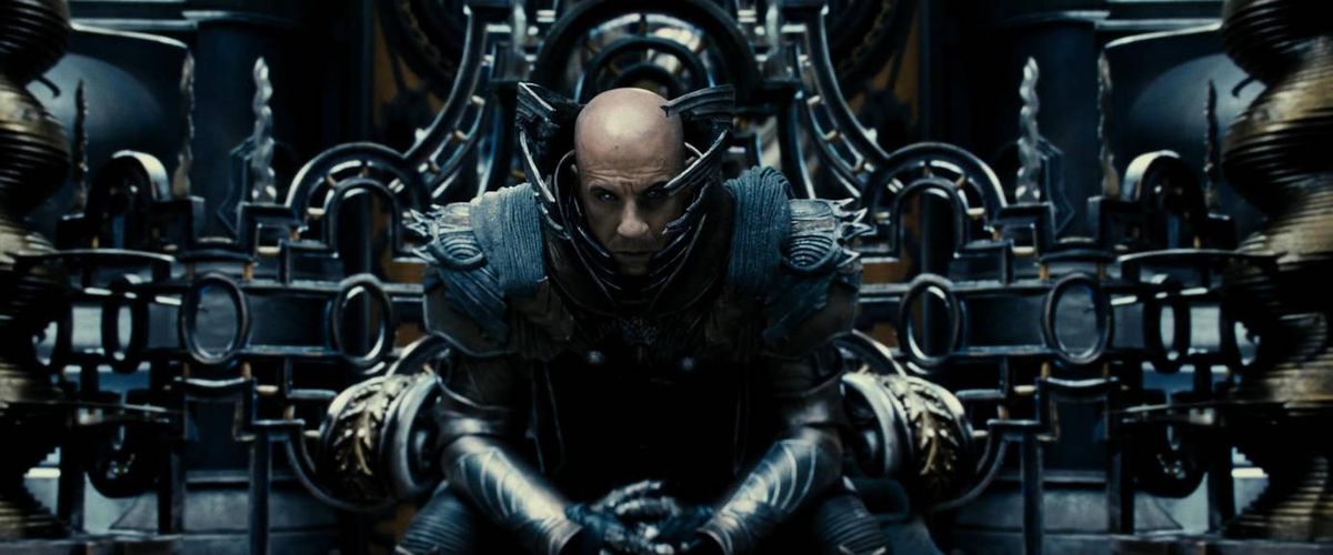 Un uomo calvo (Vin Diesel) è seduto su un trono e vestito con un'elaborata armatura metallica.