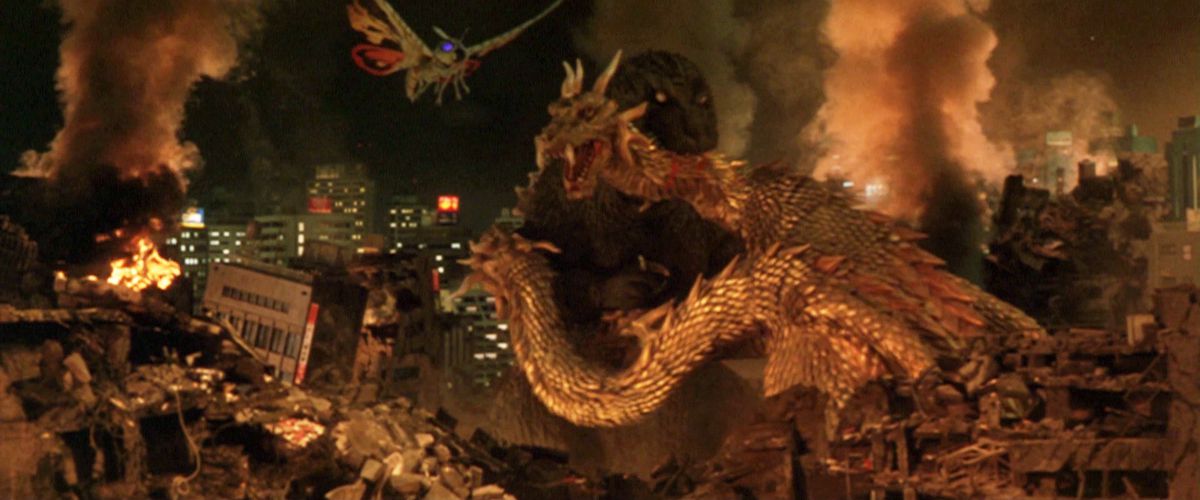 Godzilla morde il collo del re Ghidorah con una città in rovina sullo sfondo e Mothra che vola verso di loro.
