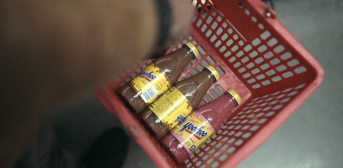 Un cesto della spesa con due bottiglie di Yoo-Hoo al cioccolato e una bottiglia di Yoo-hoo alla fragola, tenuto da un uomo dalla pelle scura con un cappotto marrone chiaro nella Stazione Undici