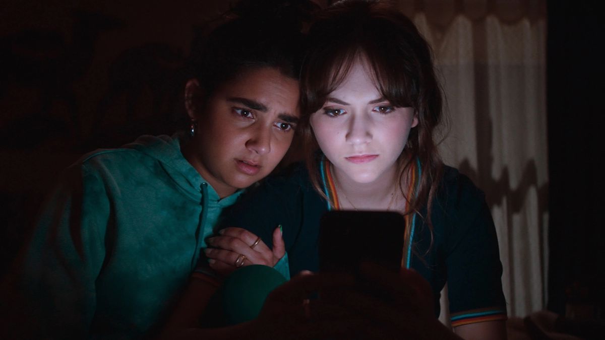 Cat Person interpreta Emilia Jones e Geraldine Viswanathan rannicchiate attorno a un dispositivo mobile, illuminate dal bagliore mentre scorrono