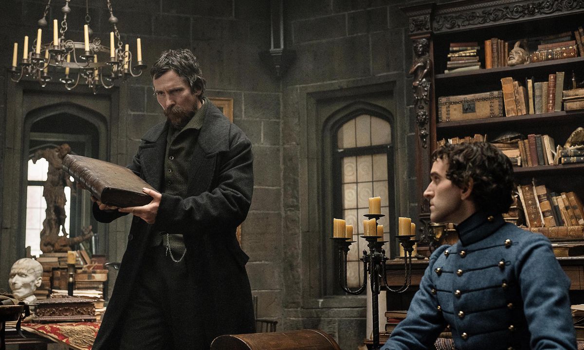 Il detective del 1830 August Landor (Christian Bale) fa una smorfia davanti a un libro in una biblioteca dell'accademia militare di West Point piena di candele mentre il cadetto Edgar Allen Poe (Harry Melling) guarda in The Pale Blue Eye