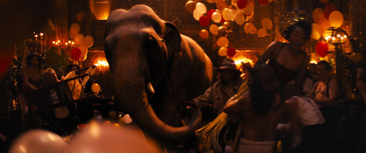 Un elefante attraversa a grandi passi il pavimento di una sala da ballo illuminata da una fioca luce rossa, spingendo tra i partecipanti alla festa e i palloncini in Babylon