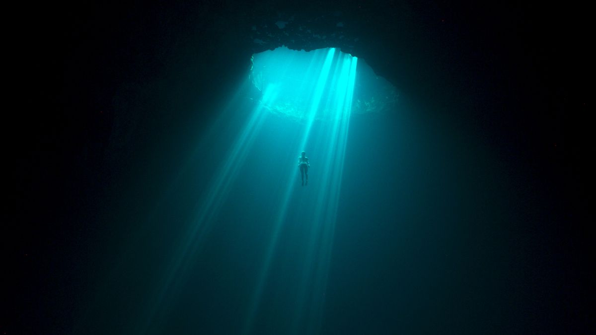 una scena oceanica profonda e oscura illuminata da un raggio di acqua turchese mentre la luce scorre attraverso l'apertura di una grotta.  un piccolo subacqueo nuota nel mezzo