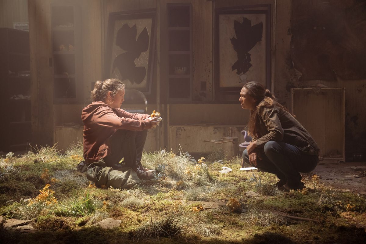 Ellie e Tess si accovacciano l'una di fronte all'altra nell'erba che ha superato un edificio decrepito mentre la luce risplende dall'alto nella serie HBO The Last of Us.