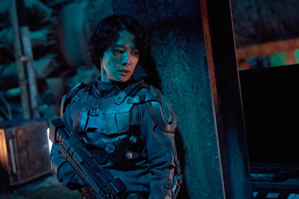 Una donna in armatura futuristica si nasconde dietro un pilastro di cemento con in mano un fucile.