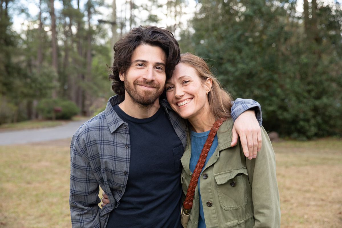 Un uomo (Jake Hoffman) con una camicia blu scuro posa per una foto con una donna sorridente (Schuyler Fisk) con una giacca verde oliva.