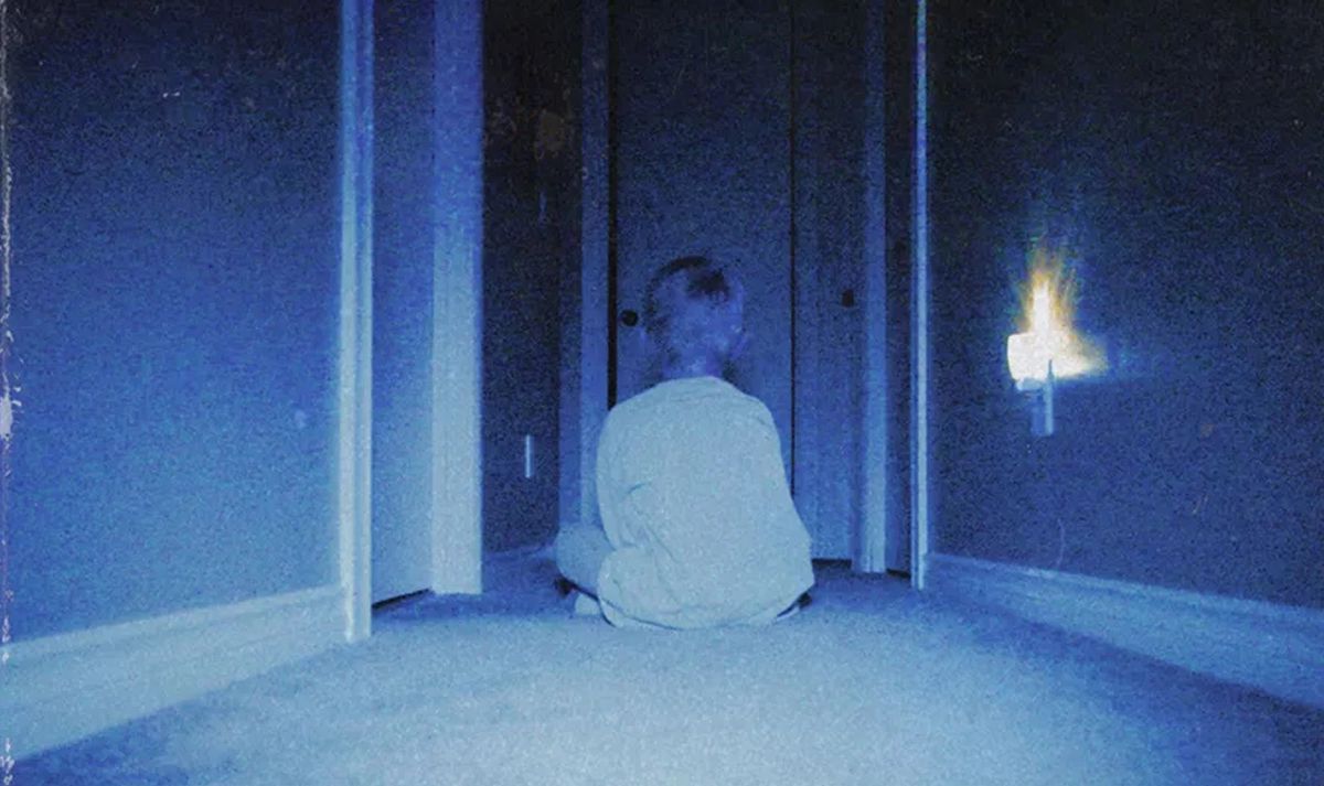 Un ragazzino siede in un buio corridoio blu con le spalle alla telecamera, di fronte a una serie di porte aperte, in una ripresa tipicamente sgranata e sfocata del film horror Skinamarink